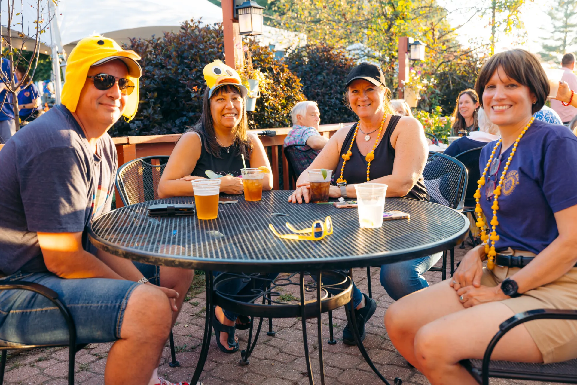 Group of four wearing yellow duck hats, necklaces, and duck-themed attire sit at a deck table during event smiling at camera